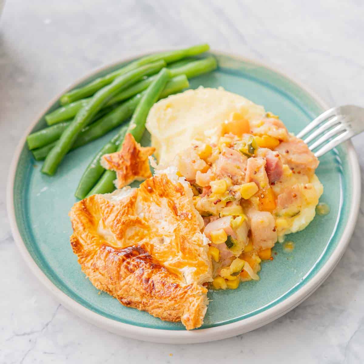 a serving of Ham Pot Pie with a side of blanched beans on a teal coloured ceramic plate and with a fork resting on the side, sitting the bench