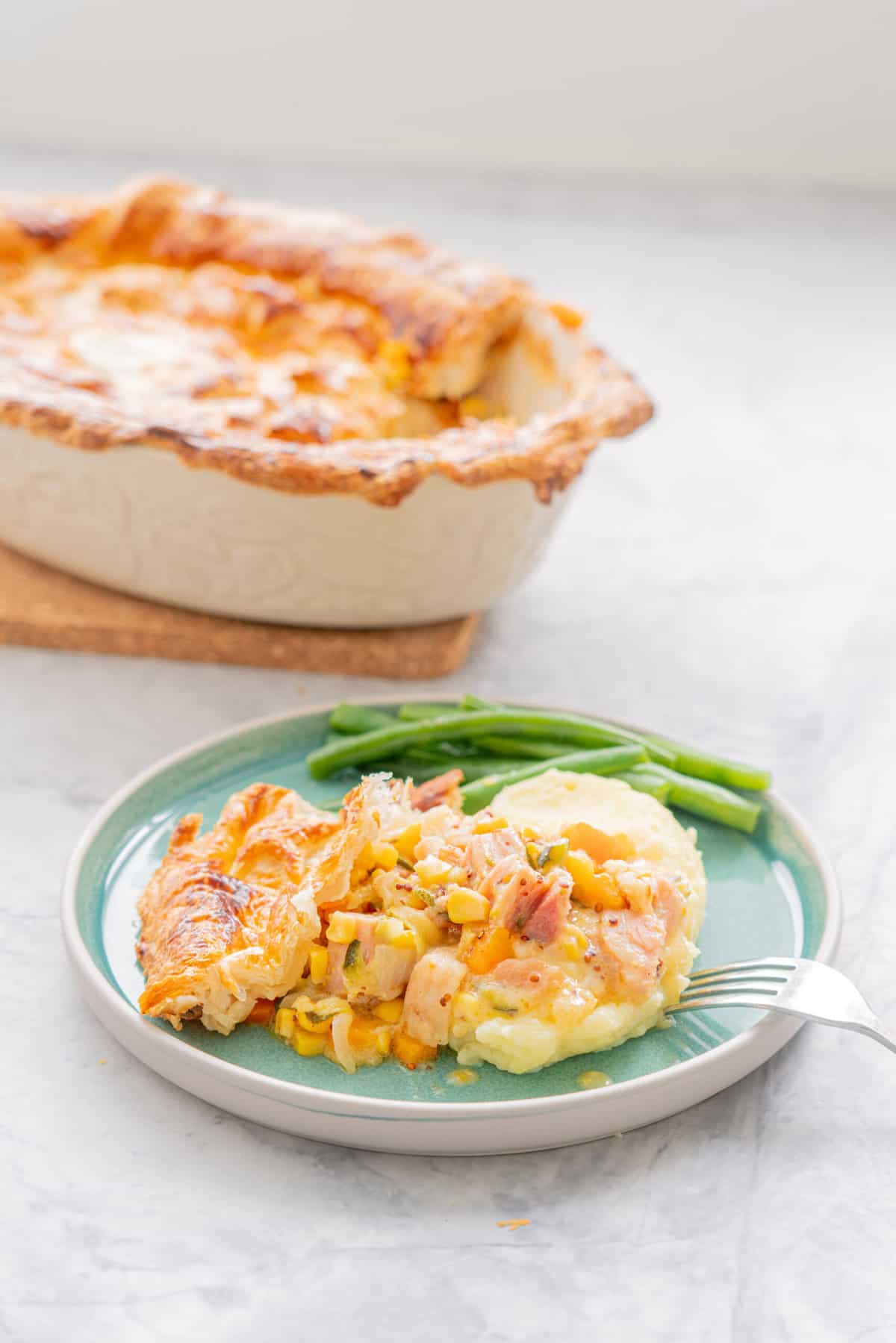 Golden brown baked ham pot pie resting on a chopping board, behind a serving with a side of beans on a teal coloured ceramic plate and a fork resting on the side