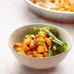 A bowl of saucy chicken, and green steamed vegetables on a marble bench top.