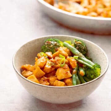 A bowl of saucy chicken, and green steamed vegetables on a marble bench top.