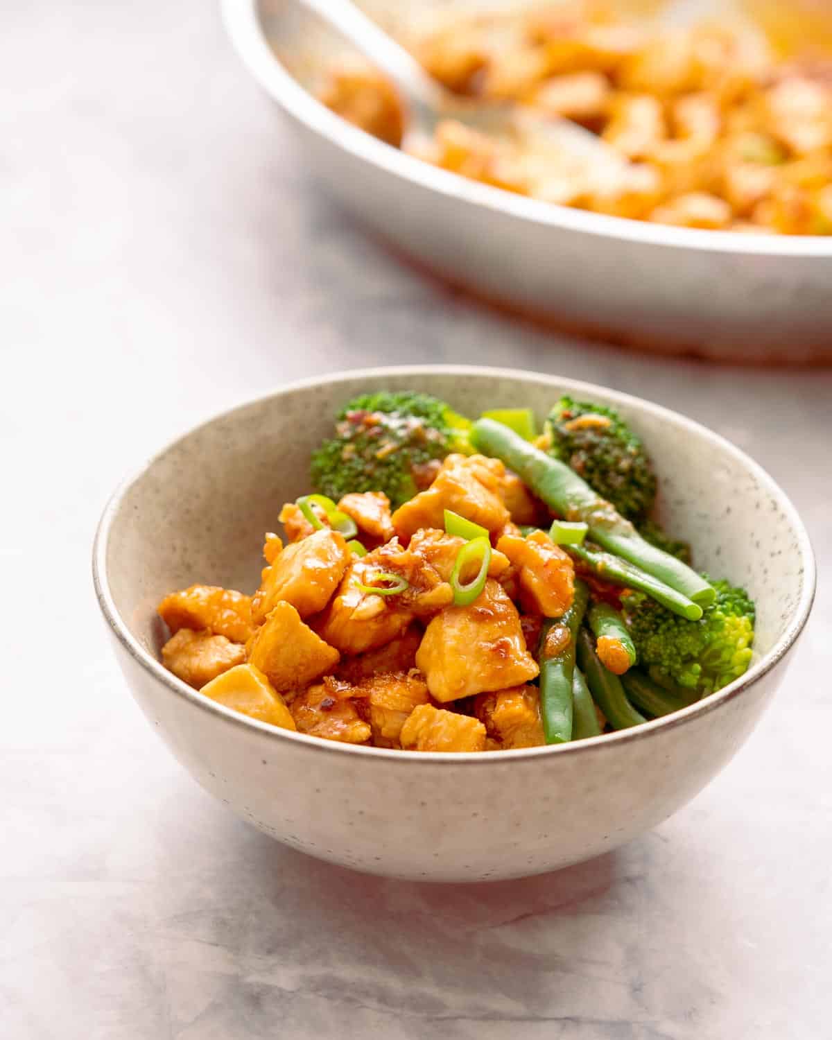 A small serving of honey garlic chicken with a side of blanched beans and broccoli sitting on a bench with the frying pan of the dish in the background