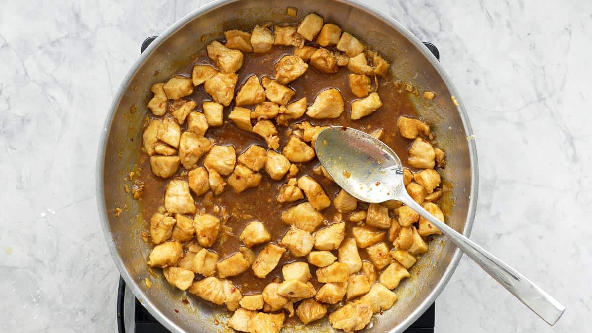 Diced chicken cooking on the element with the honey garlic sauce added and a spoon resting on the side