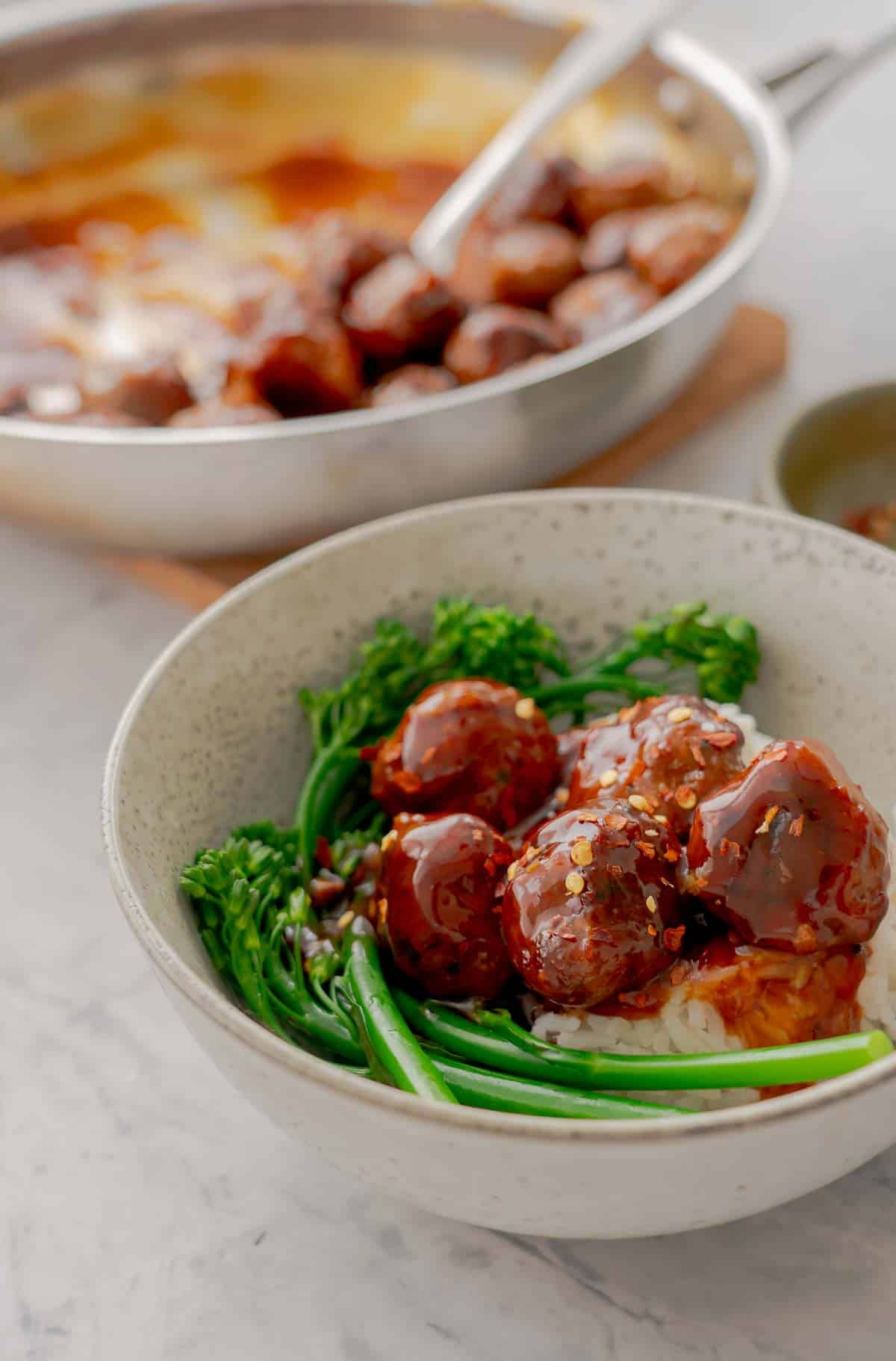 A bowl with meatballs in a sticky sauce, steamed brococlini and rice.