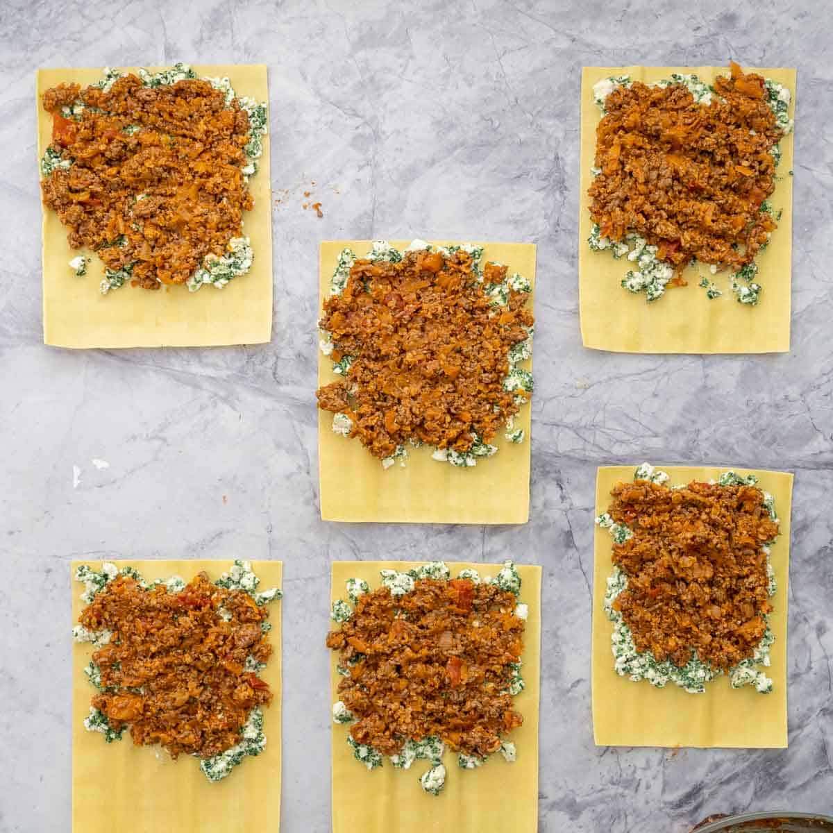 Fresh lasagne sheets laid out on the bench with each sheet spread with a layer of spinach and cottage cheese mixture and topped with a layer of meat sauce.