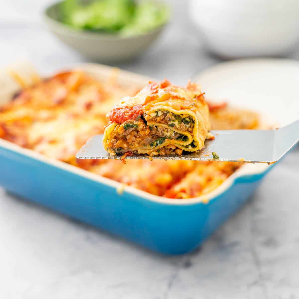 Baked Lasagna roll ups in a blue oven dish sitting on the bench with a green salad in the background and a spatula full of lasagna raised above the dish