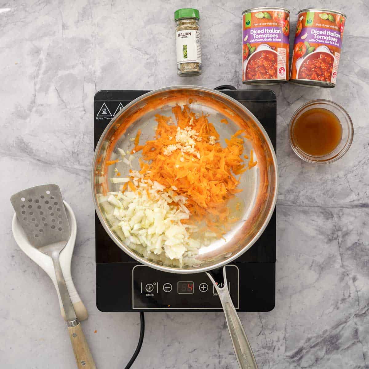 fry pan on a bench top cooker, with the onion, garlic and carrots sautéing next to two cans of diced tomatoes and a cup of broth and a spatula.