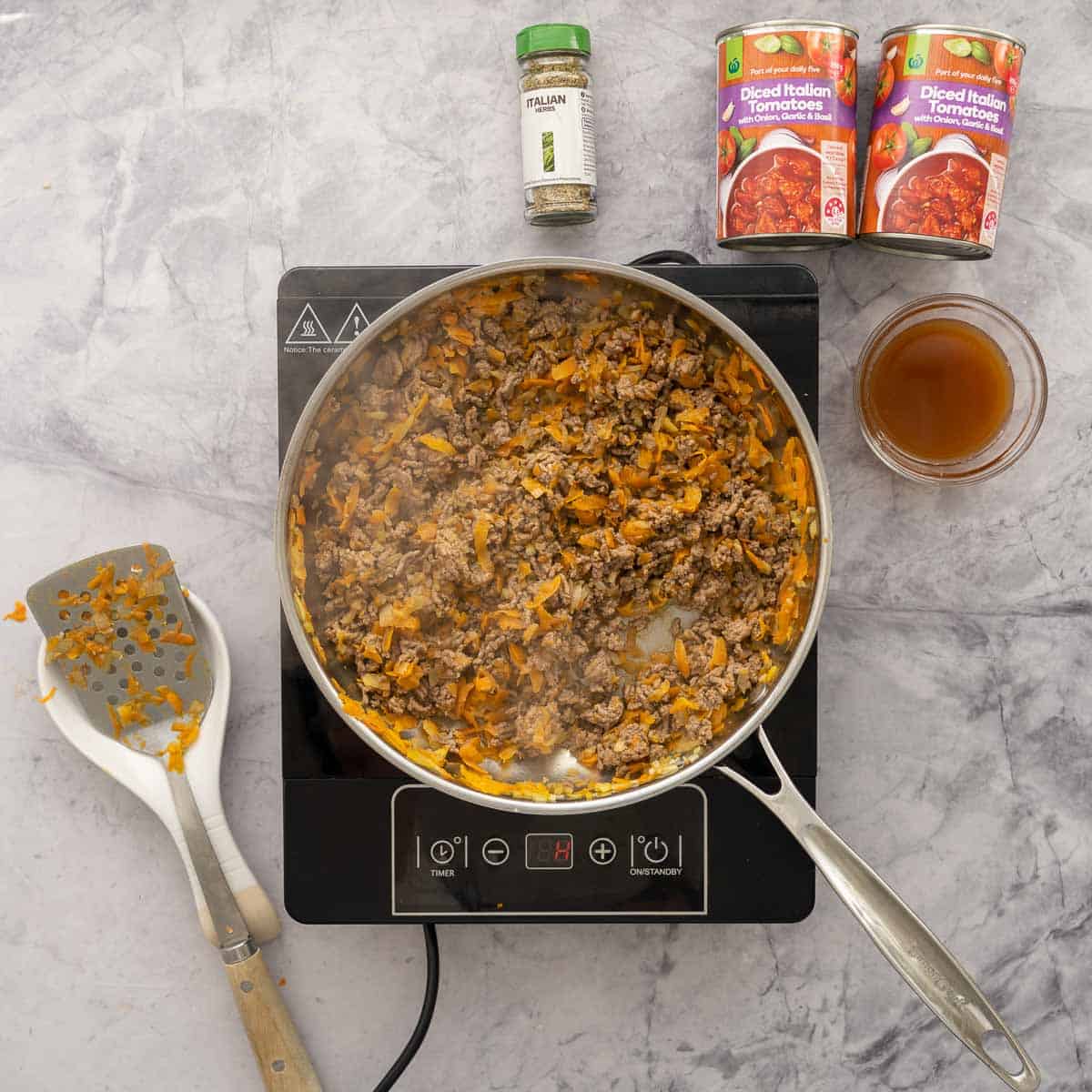 Beef mince added to the pan which is sitting on a bench top cooker next to two cans of diced tomatoes and a half cup of broth and spatula.
