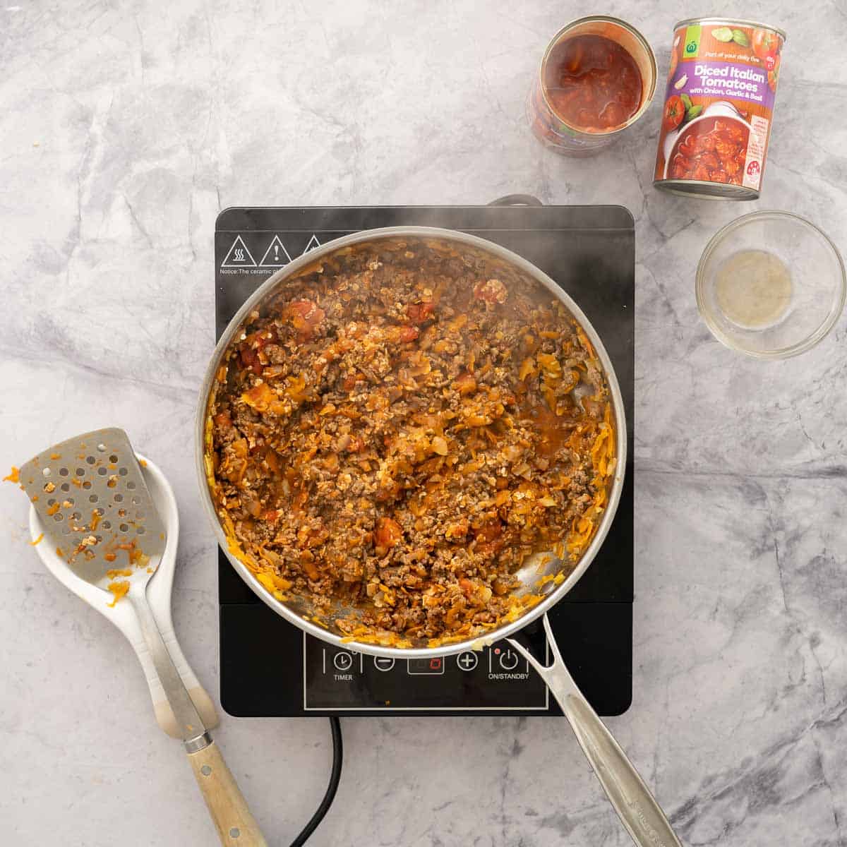 Minced meat mixture cooking on a stove top cooker with a emptied cans of diced tomatoes, a glass bowl and a spatula