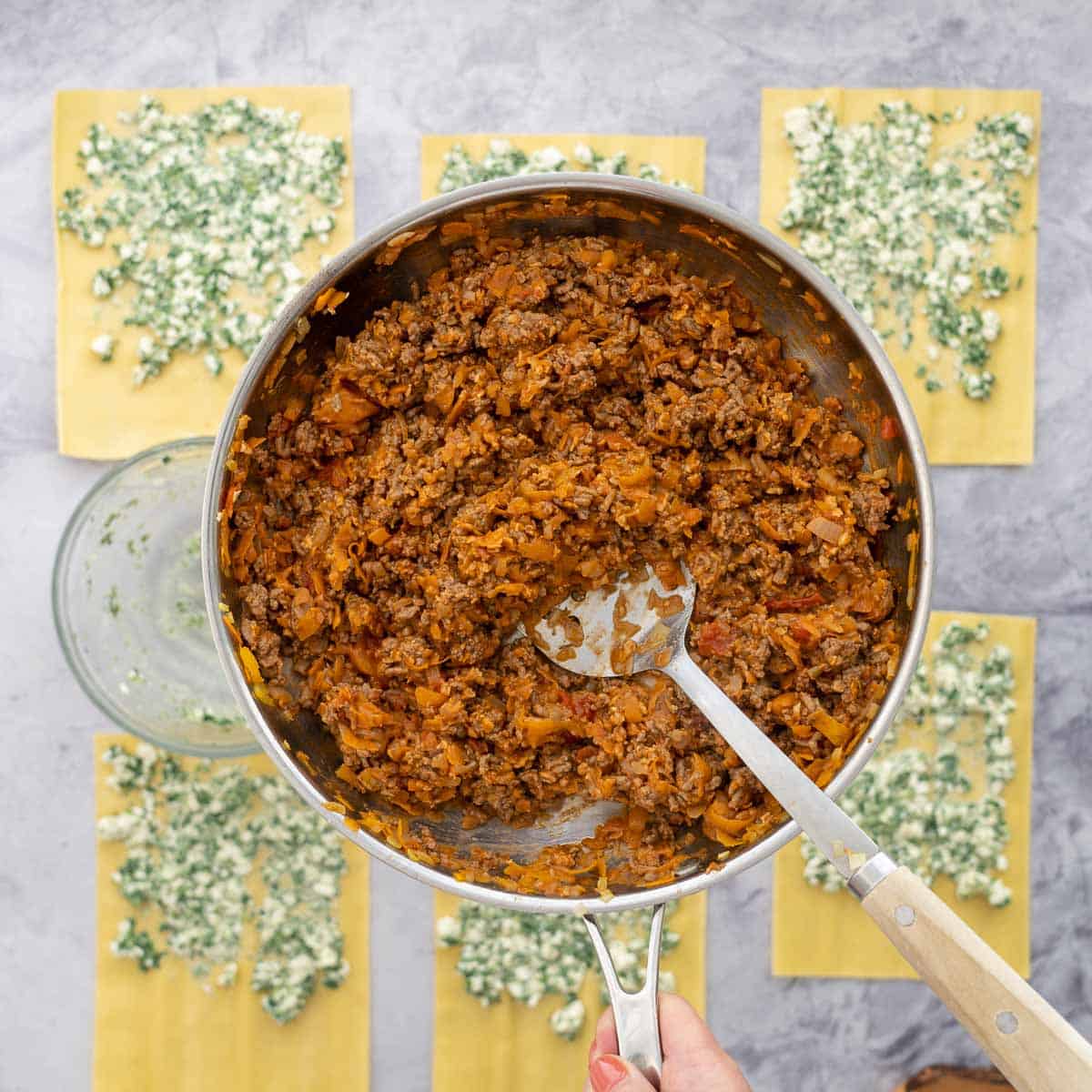 Fresh lasagne sheets laid out on the bench with each sheet spread with a layer of spinach and cottage cheese mixture and the frying pan of meat mixture held above them,