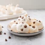 A plateful of stacked meringue cookies with a scattering of chocolate drops on the bench top and the remaining batch in the background.