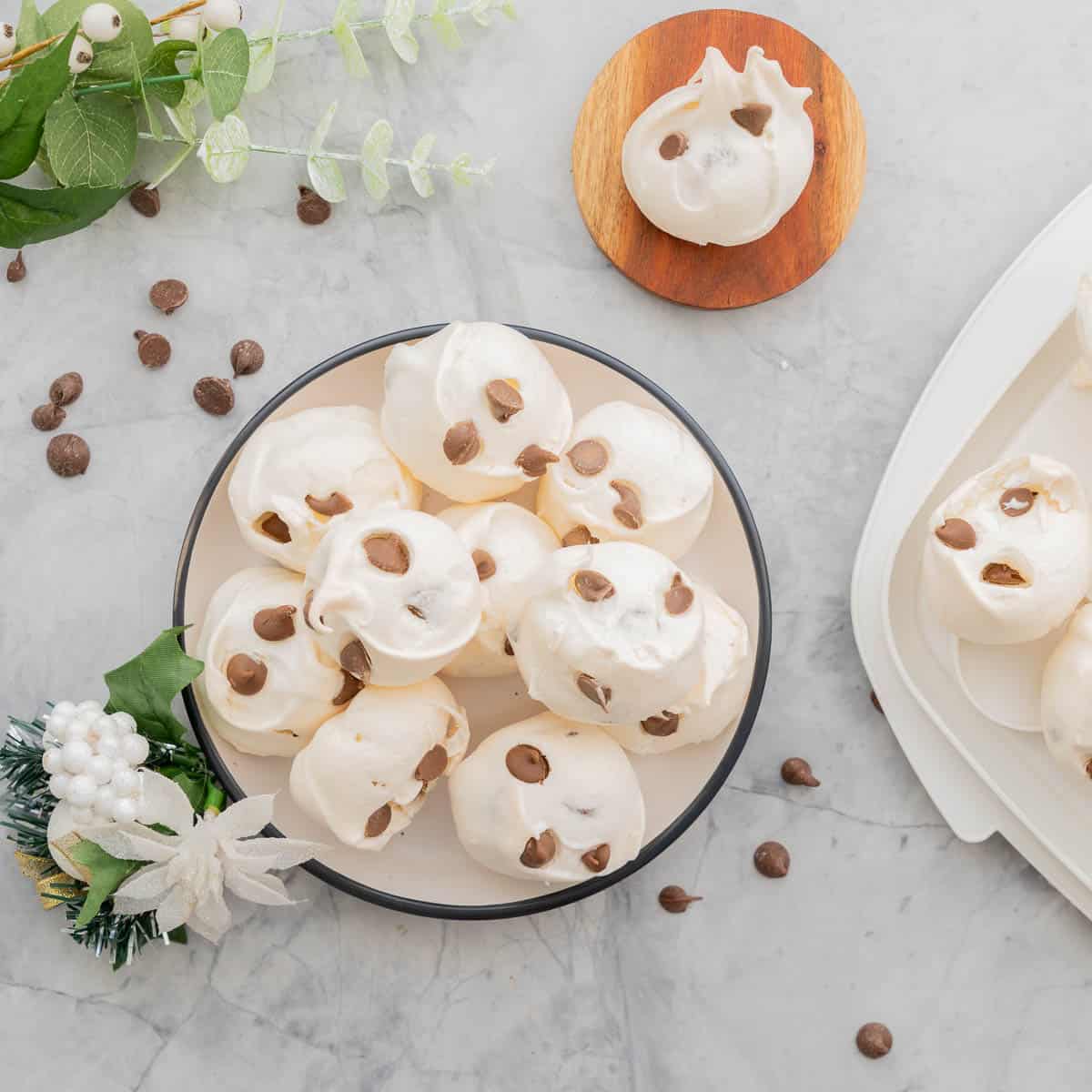 Meringue cookies stacked on a round serving dish on the bench next to scattered chocolate drops and a green and white floral Christmas decoration.