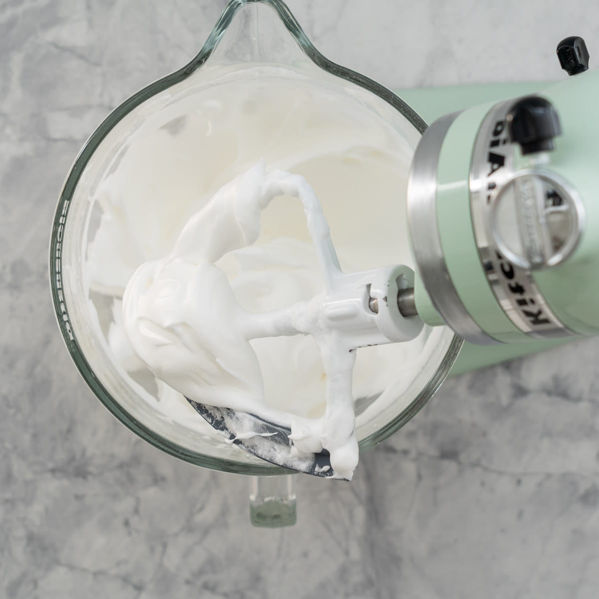 A stand up mixer sitting on the bench with whipped meringue cookies mixture with the beaters lifted showing soft peaks