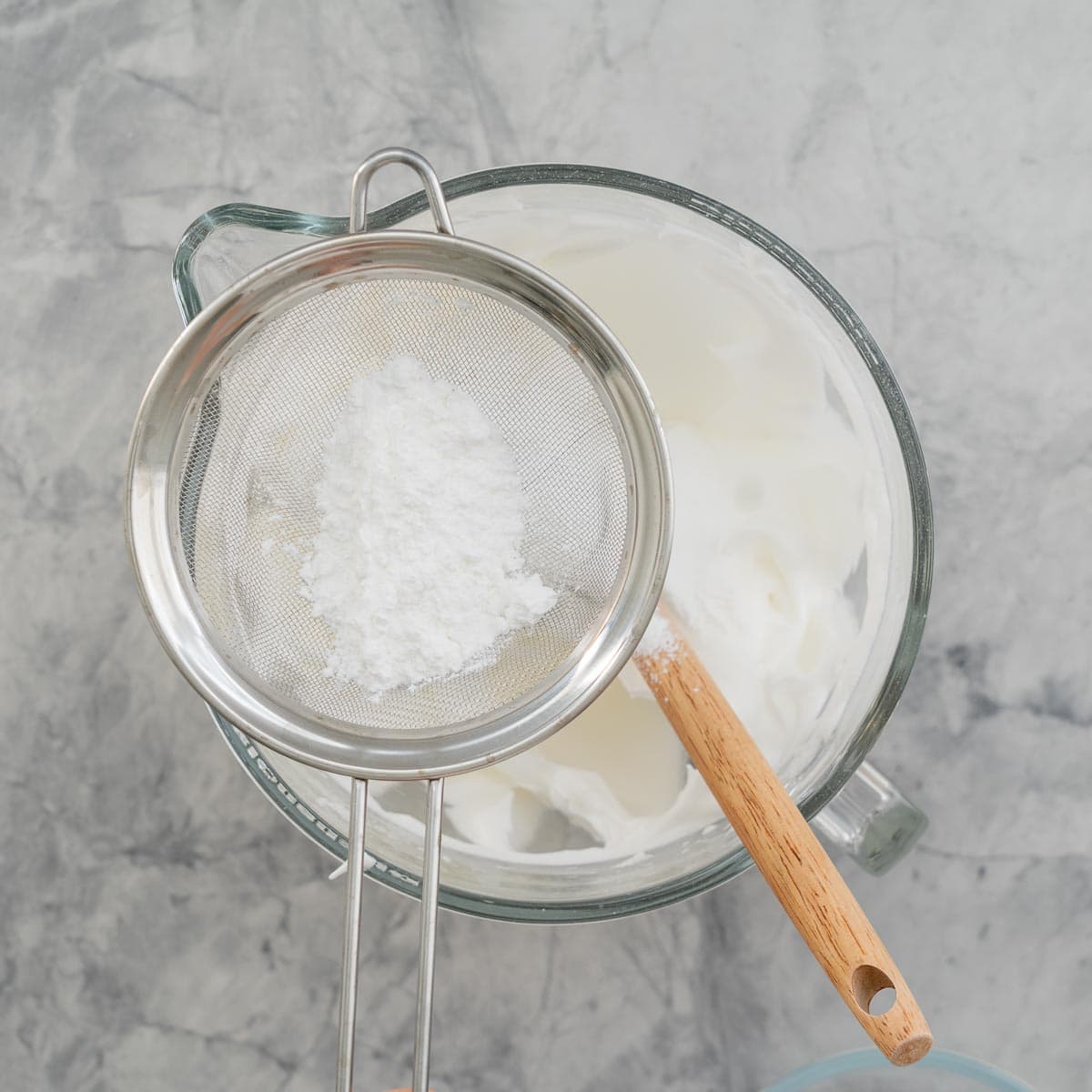 A stand up mixer bowl sitting on the bench with whipped egg whites and a sieve full of icing sugar above it.