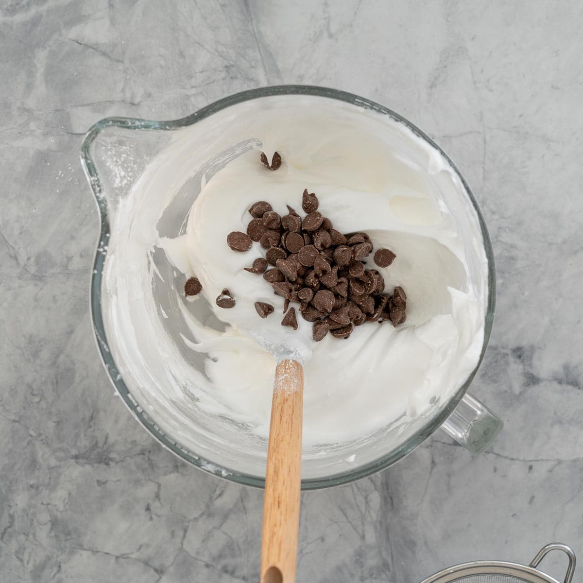 Whipped Meringue cookie mixture with a sprinkling of chocolate chips in a glass stand up mixer bowl resting on the bench.