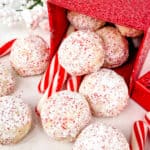 Snowball cookies falling out of a red present box onto benchtop with candy canes scattered around.