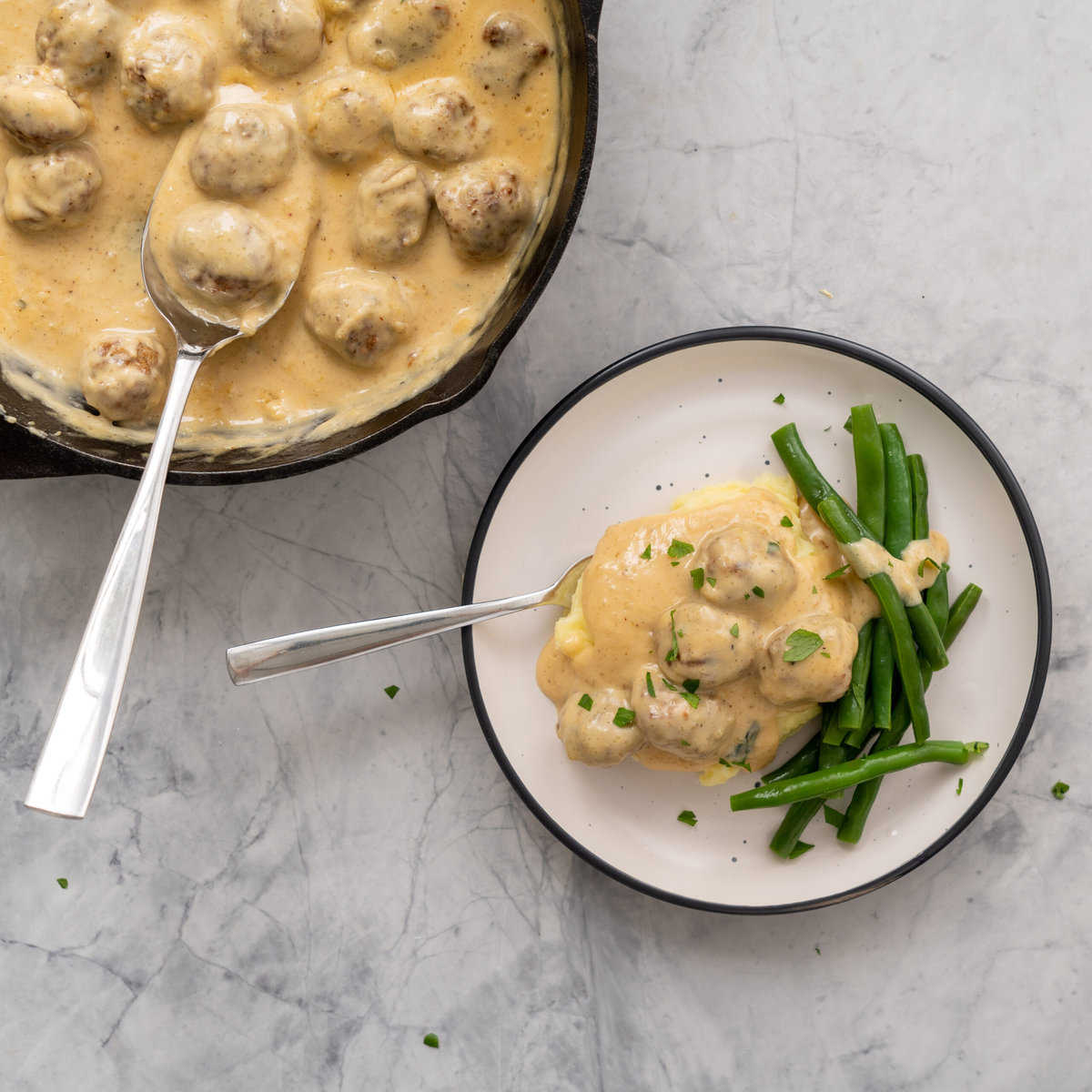 A large spoonful of Sweedish meatballs served over the top of mashed potatoes and side of blanched beans with a sprinkling of parsley.