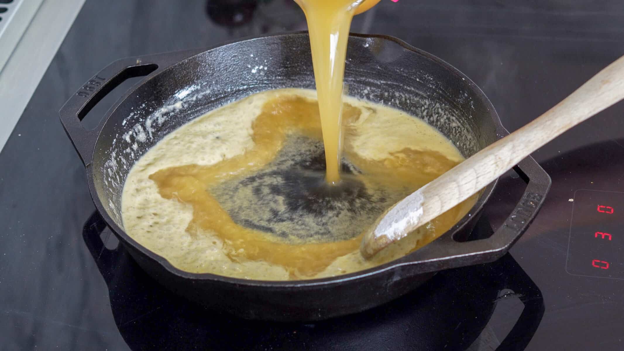 Beef stock being stirred into the cast iron pan of melted butter and flour by a wooden spoon.