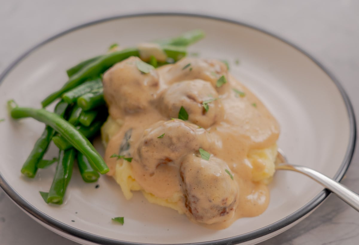 A large spoonful of Sweedish meatballs served over the top of mashed potatoes and side of blanched beans with a sprinkling of parsley.