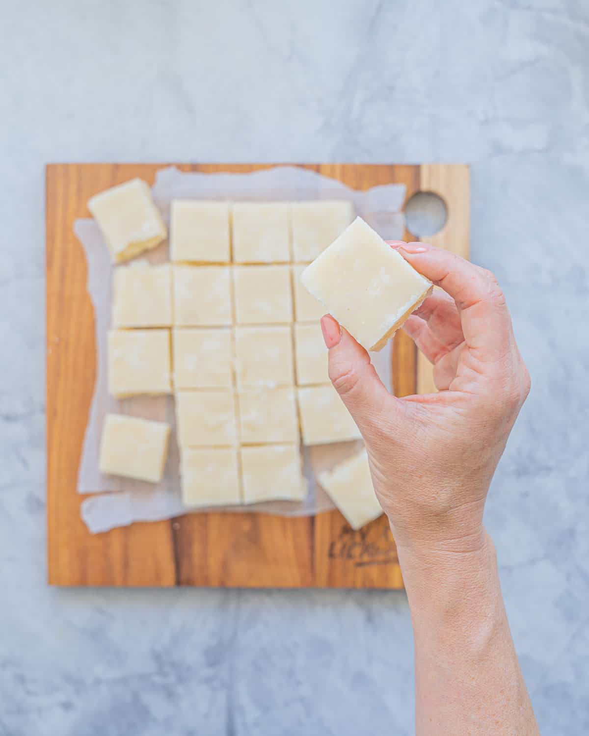 A hand with tan nail polish holding up a slice of vanilla fudge above the rest of the batch which is sitting on the bench below