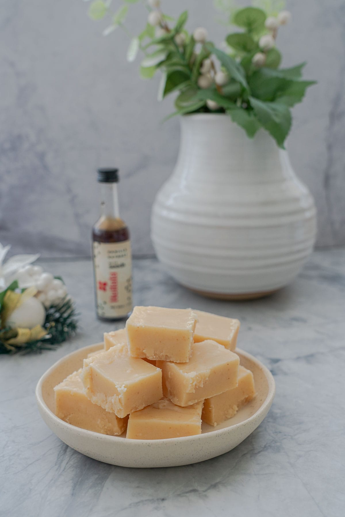 Slices of vanilla fudge stacked in a small serving dish which is resting on the bench next to a bottle of vanilla essence and a ceramic vase full of green leaves.