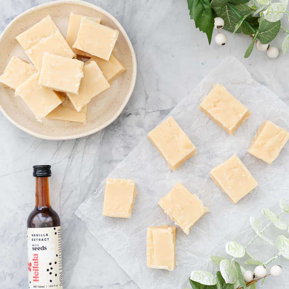 Slices of fudge resting a sheet of crinkled baking paper next to a serving dish of stacked slices and a small bottle of Heilala vanilla extract and christmas holly
