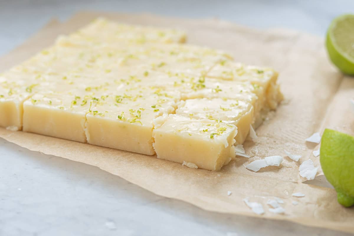 Sliced Vegan Fudge sitting on the bench with a sprinkling of lime zest on top and a scattering of coconut flakes next to two lime haves.