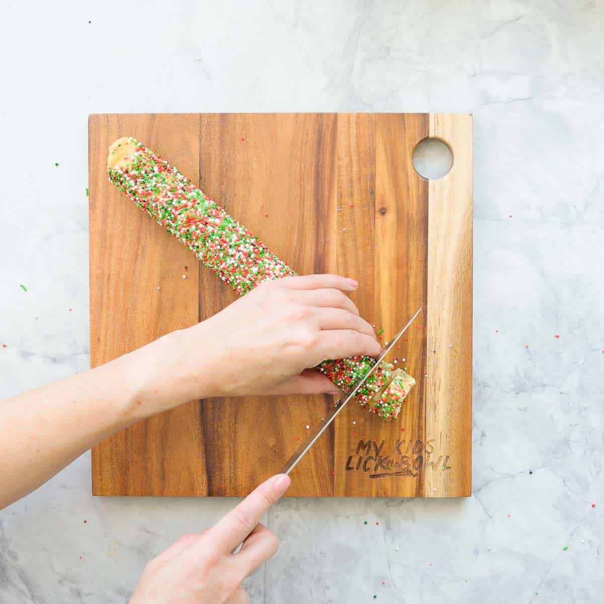 One cookie dough log covered in Christmas coloured sprinkles being sliced into one cm wide disks on a wooden chopping board