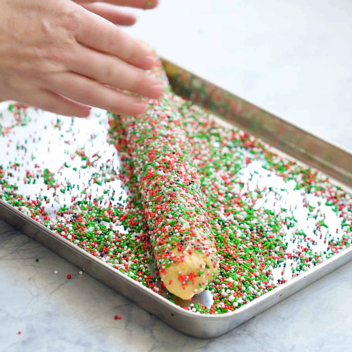 Unwrapped chilled cookie dough log being rolled in a large baking tray of Christmas coloured sprinkles.
