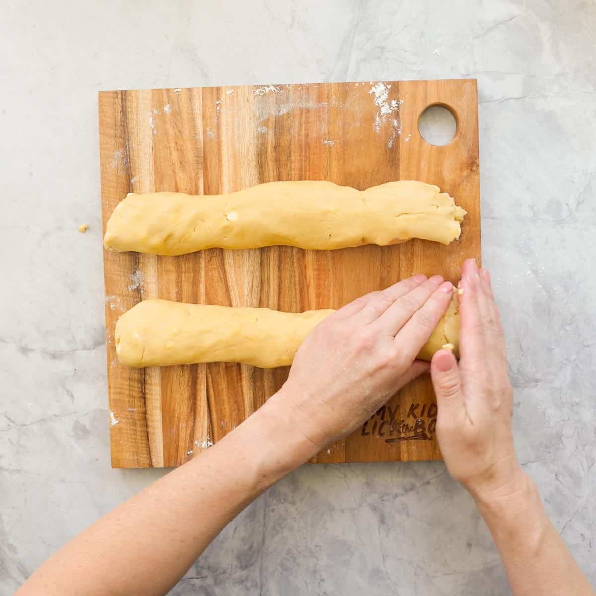 Hands shaping and moulding the cookie dough into two logs on a wooden chopping board.
