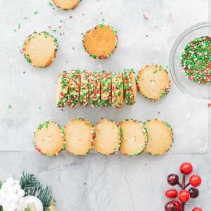 Baked disks of Christmas Cookies resting on the bench next to a glass bowl of green, red and white sprinkles.