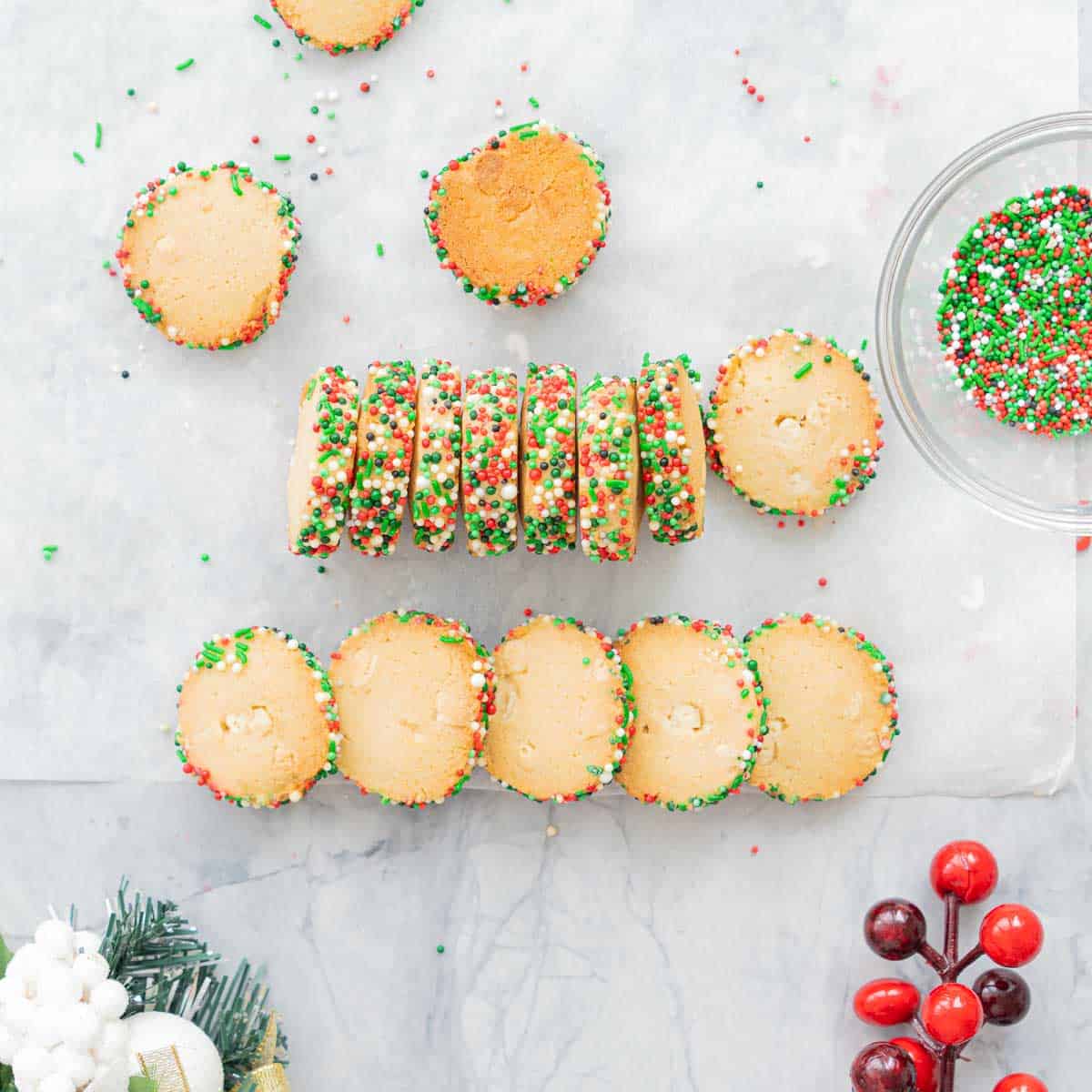 Baked disks of Christmas Cookies resting on the bench next to a glass bowl of green, red and white sprinkles