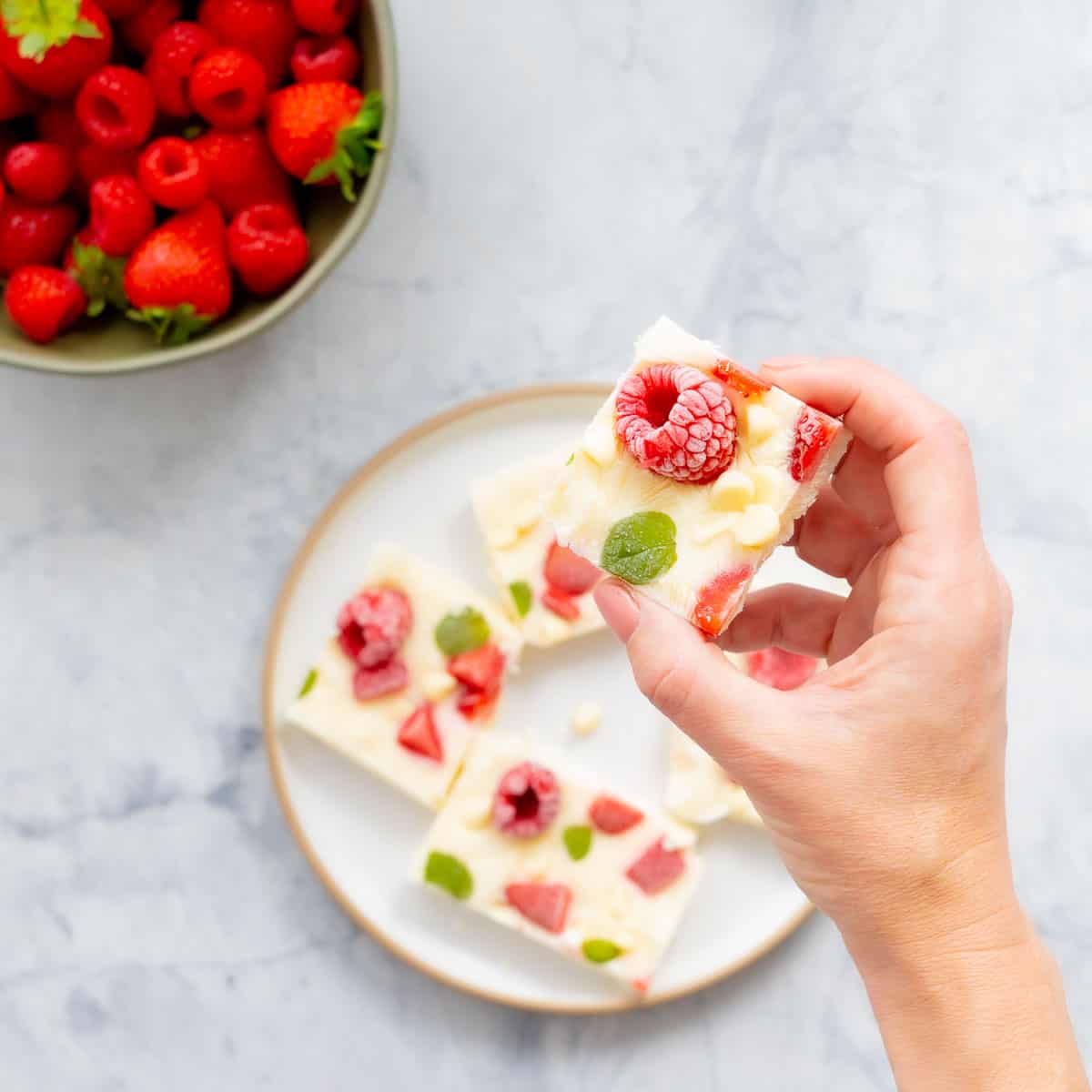 A hand holding up a slice of yoghurt bark above a plateful below which is sitting on the bench.