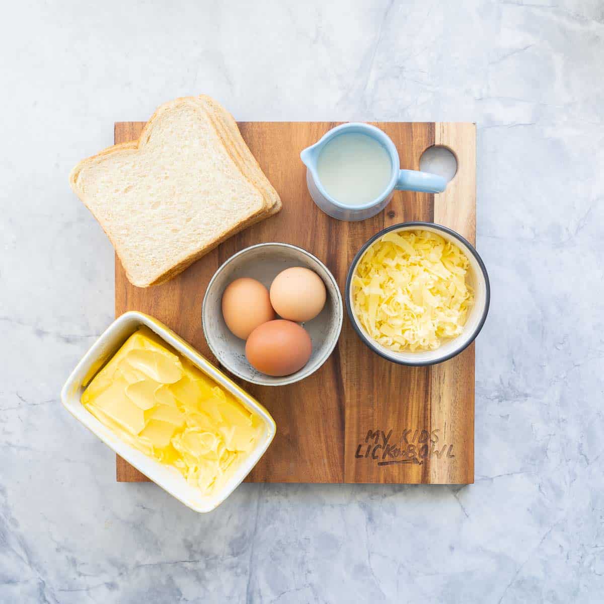 Chopping board with three slices of bread, small jug of milk, grated cheese, three eggs and butter dish.