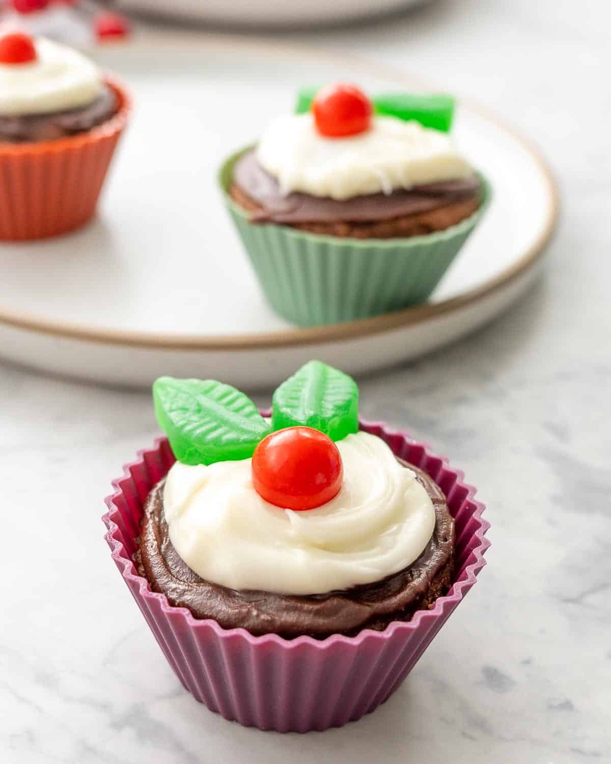 A cupcake in a burgandy cupcake liner decorated with frosting and candy to look like a figgy pudding.