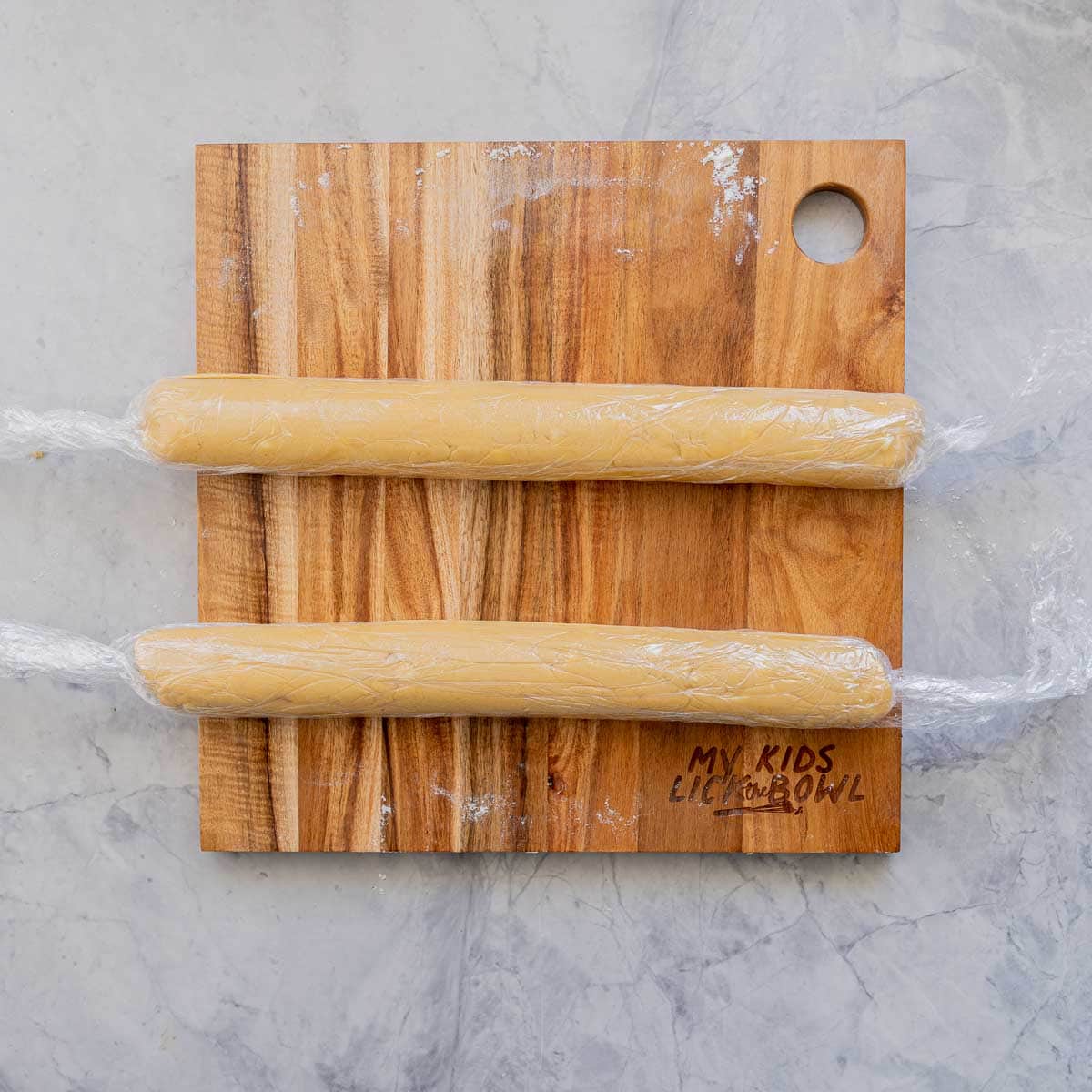 Two chilled cookie dough logs wrapped it in cling film resting on a wood chopping board on the bench