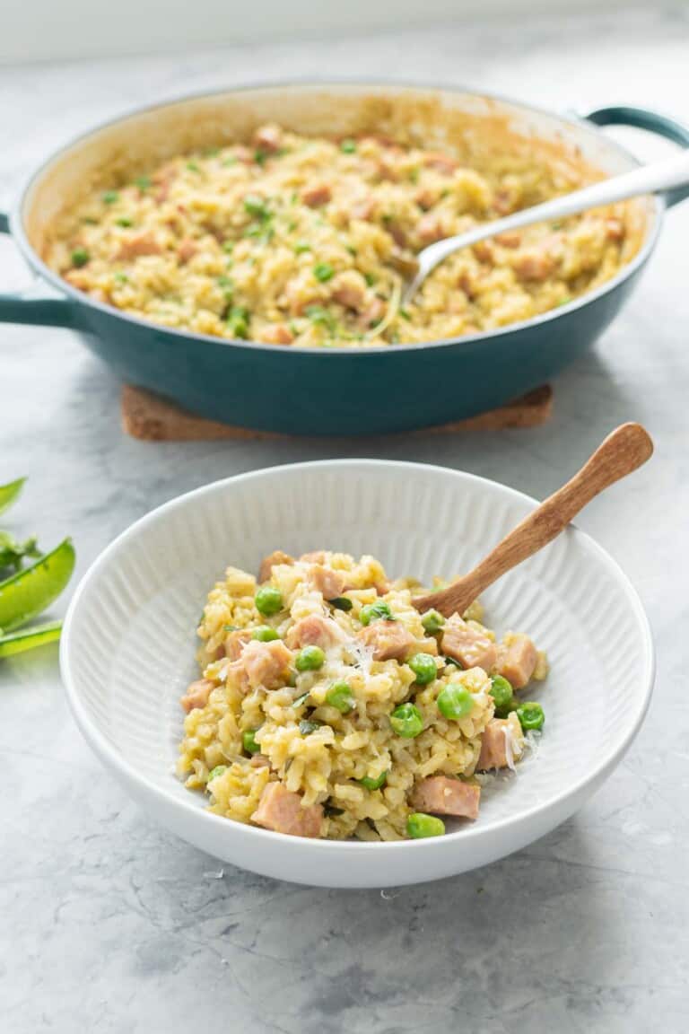 A bowl of kakhki green risotto studded with green peas and chunks of pink ham in a ceramic bowl with wooden spoon.
