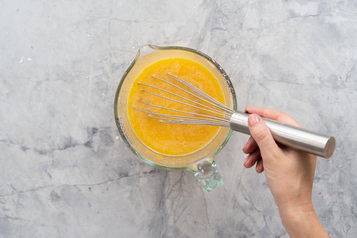 A hand whisking together the egg mixture with lemon juice in a glass jug on the bench