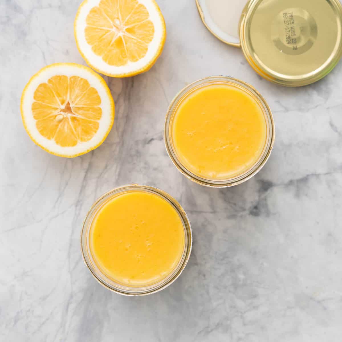 Two glass jars of lemon butter resting on the bench next to a halved lemon and 2 gold lids