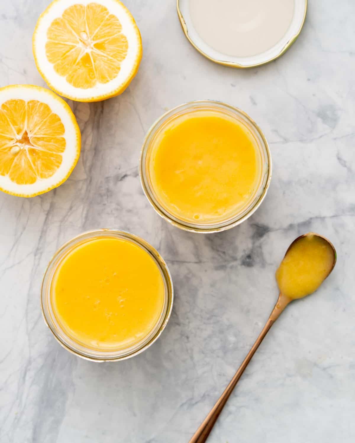 Two glass jars of lemon butter resting on the bench next to a halved lemon and a brass spoon resting on the bench with a spoonful ofthelemon butter