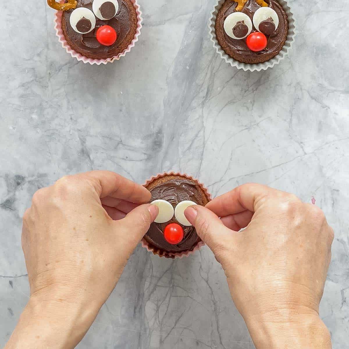 A chocolate frosted cupcake with a red candy placed in the centre and two white chocolate buttons placed above it