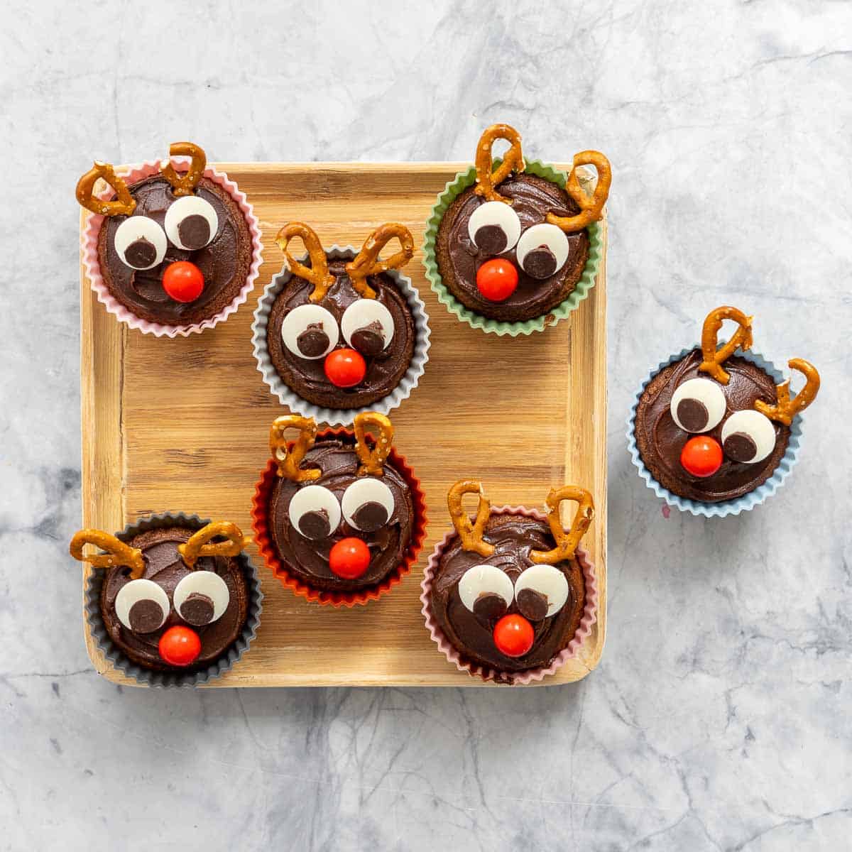 A square bamboo tray with seven chocolate cupcakes decorated to look like reindeer.