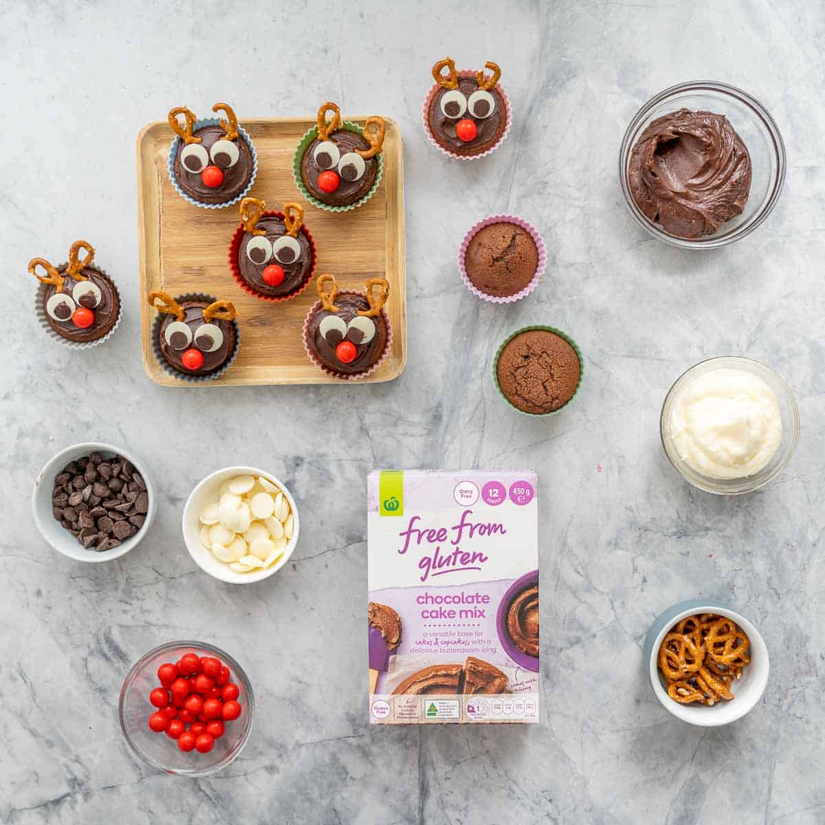 Reindeer cupcakes, a packet of chocolate cake mix, bowls of frosting, pretzels, candy and chocolate buttons on a marble bench top.