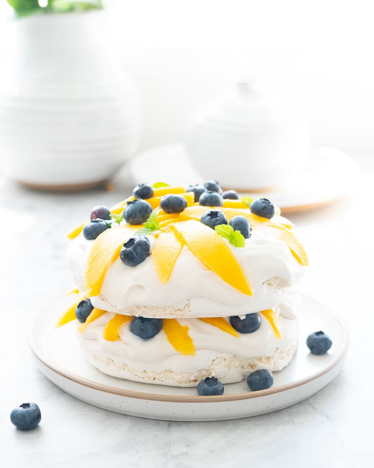 A two layer pavlova topped and filled with whipped cream, sliced mango, blueberries and mint leaves