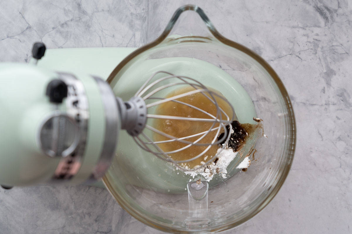 Aquafaba, vanilla extract and cream of tartar in the glass mixing bowl of a stand mixer.