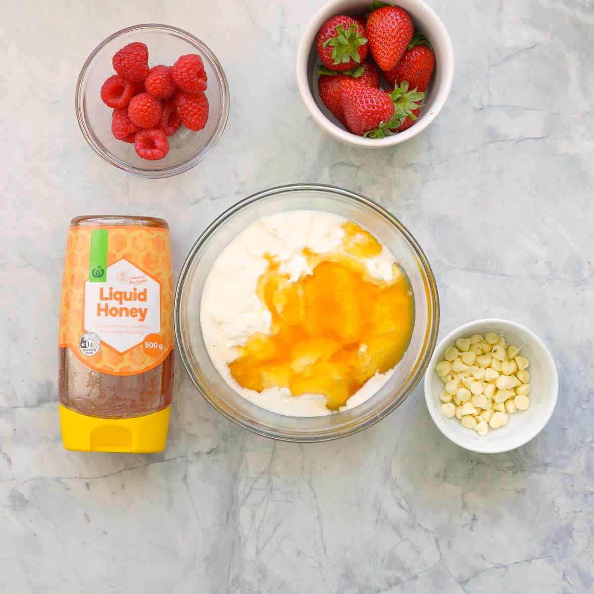 A small glass mixing bowl with yogurt, vanilla extract and honey sitting next to a bottle of liquid honey and ramekins of raspberries, strawberries and white chocolate drops on the bench