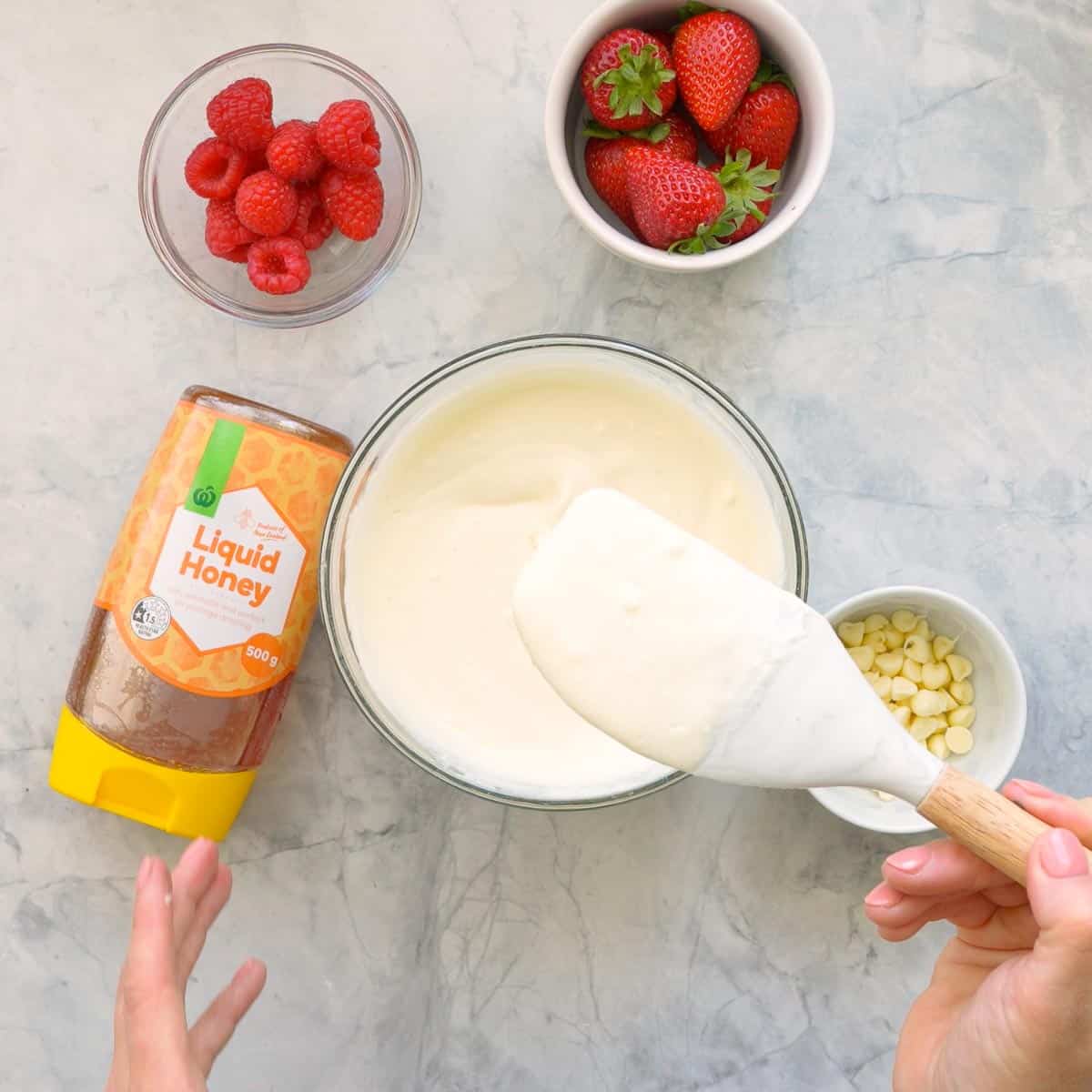 A small glass mixing bowl with yogurt, vanilla extract and honey stirred together with a spatula which issitting next to a bottle of liquid honey and ramekins of raspberries, strawberries and white chocolate drops on the bench