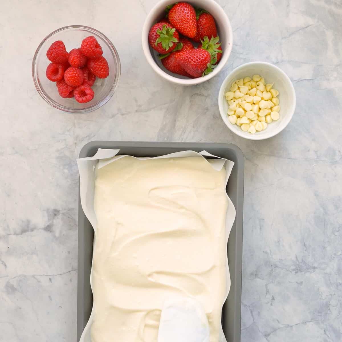 A lined baking tray with the yoghurt mixture spread out evenly which is sitting next to ramekins of white chocolate, raspberries and strawberries