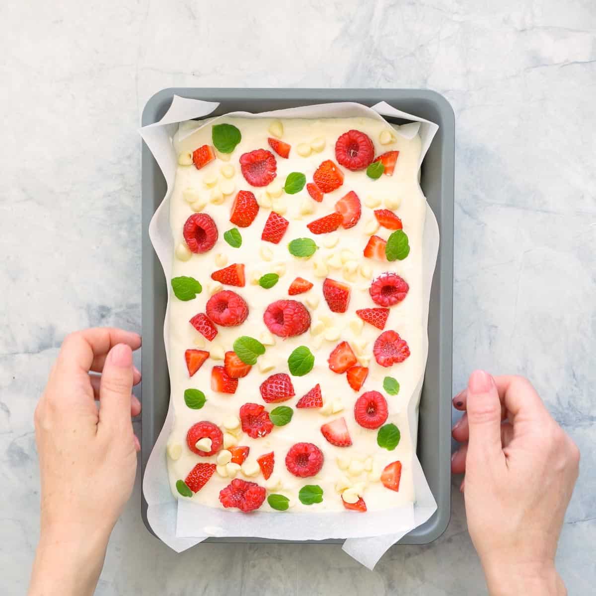 A lined tray of yoghurt bark which is scattered with sliced strawberries, raspberries and baby mint leaves scattered over the top with two hands resting either side of it on the bench.