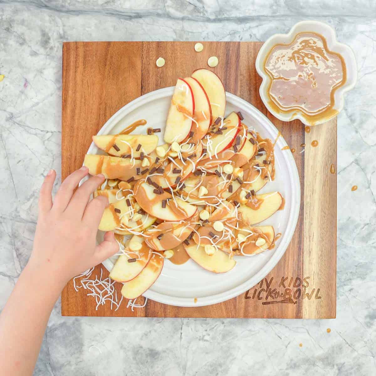 A child's hand reaching to take apple nachos off a serving platter.