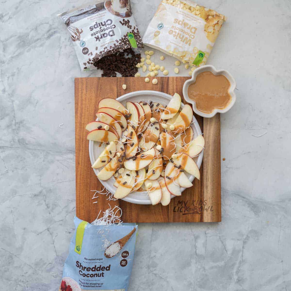 Apple slices arranged on a round white platter drizzled with caramel and scattered with chocolate chips and shredded coconut.